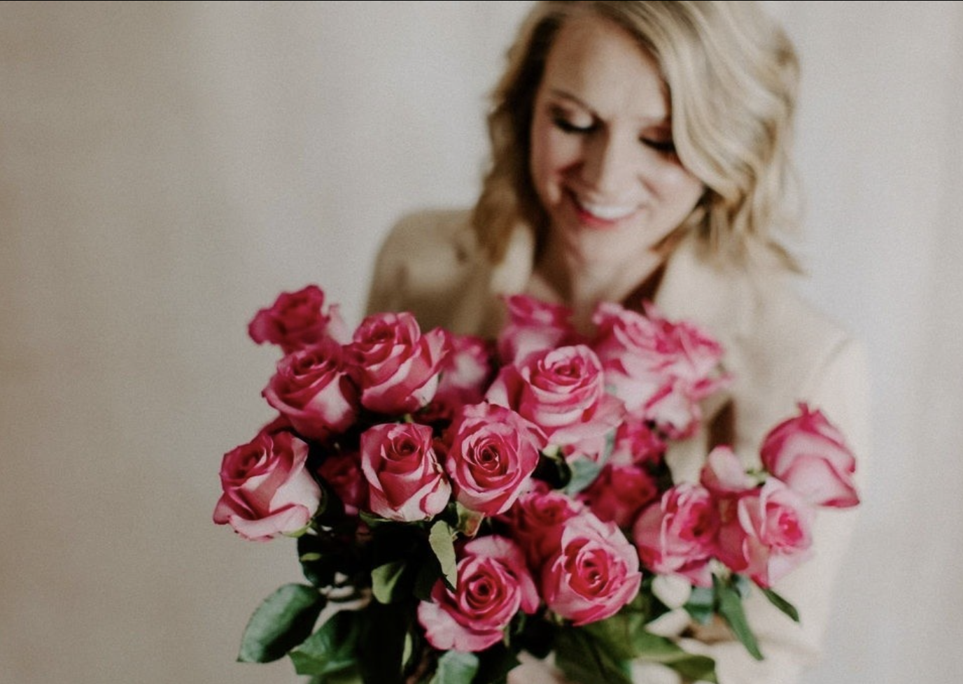 blonde girl in beige blazer smiling down at pink roses